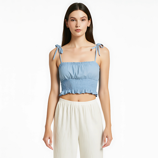 Woman wearing sky blue ruched cami top with tie straps, smocked waist, and ruffle hem; styled with white pants.