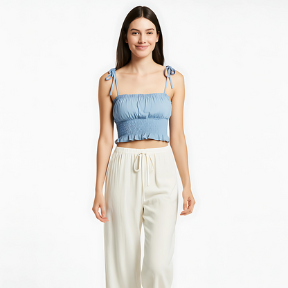 Sky blue ruched cami with bow-tied straps and ruffle hem, perfect for summer and resort wear, paired with cream-colored trousers.